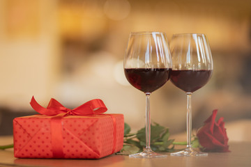 Wine glasses, present and roses on the wooden table in front of beige warm background. Love and holiday concept