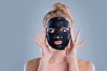 girl with naked shoulders posing at grey background with facial mask black, skin care concept, beauty photo, skin treatment, hydrating skin mask, smiling.