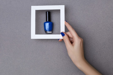 Female hand with manicure in trendy classic blue color holding white frame above a bottle of nail polish on the grey background