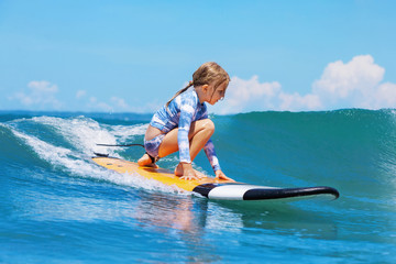 Happy baby girl - young surfer ride on surfboard with fun on sea waves. Active family lifestyle, kids outdoor water sport lessons and swimming activity in surf camp. Beach summer vacation with child.
