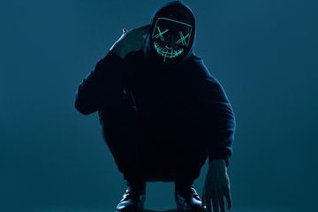 Anonymous man in black hoodie hiding his face behind a neon mask