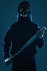 Anonymous criminal man with baseball bat in a black hoodie and neon mask