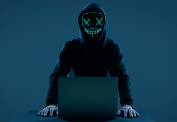 Anonymous man in a black hoodie and neon mask hacking into a computer