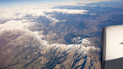 view of sky, snowy mountains, and airplane engine through window while flying