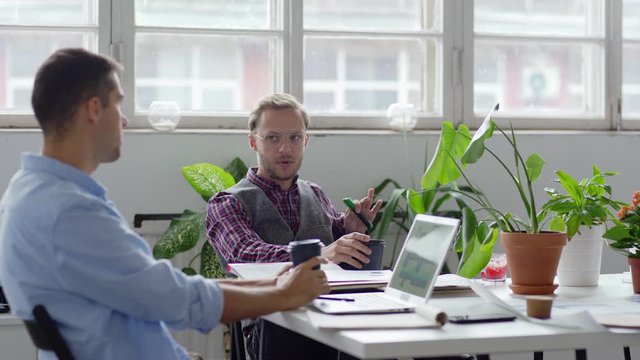 Tracking medium shot of two young entrepreneurs sitting at desk in office and talking over coffee. Man drinking takeaway coffee from disposable paper cups. Open laptop and potted flowers on table