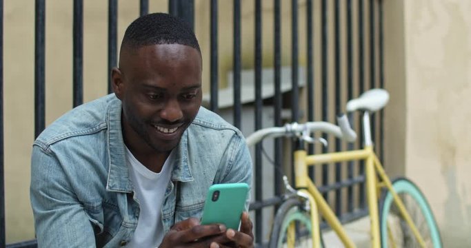 Cheerful African American Young Man Using his Smartphone for Typing SMS, Looking Excited and Laughing while Sitting near Stylish Bike with Old Metal Fance at the Background.
