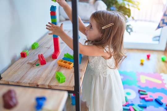 Adorable blonde toddler playing with building blocks around lots of toys at kindergarten