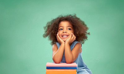 childhood, school and education concept - happy smiling little african american girl with pile of books over green chalk board background