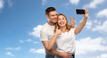 relationships, technology and people concept - happy couple in white t-shirts taking selfie smartphone and showing peace by over blue sky and clouds background