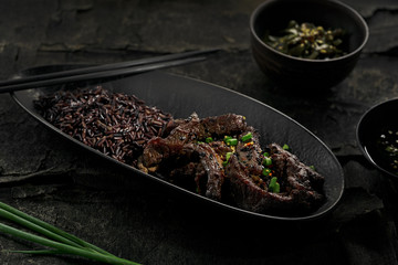 Sticky Asian beef stir fry in salty sweet sauce with black rice in a black clay plate.