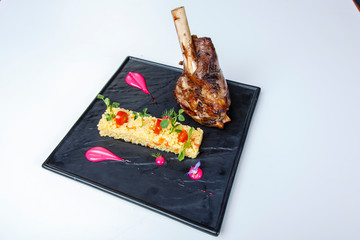 Baked pork knuckle with rice on a square black plate. Rice in the form of a bar, decorated with greens and tomatoes. 