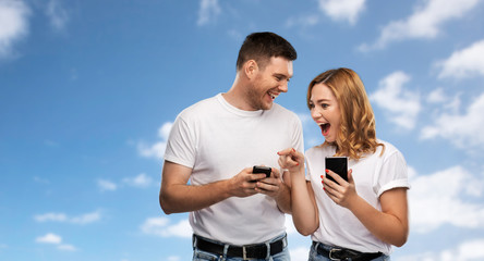 technology and people concept - happy couple in white t-shirts with smartphones over blue sky and clouds background