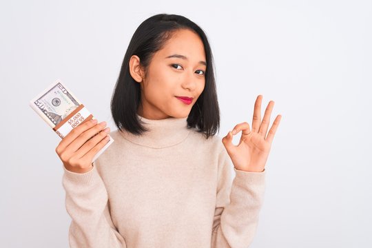 Young beautiful chinese woman holding dollars standing over isolated white background doing ok sign with fingers, excellent symbol