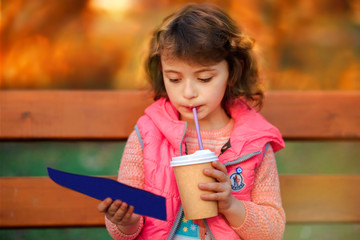 Little cute 6 years old girl in pink vest some text in sunset time in park. Child drinks drink from paper cup with pipe outdoors. Space for text. Natural light. Education for children. Self education