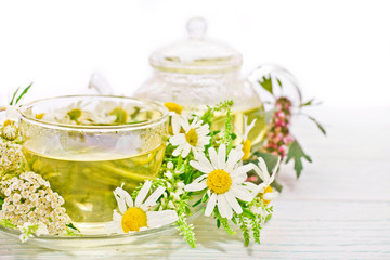 Herbal tea in a glass cup and teapot with fresh herbs on white wooden background.