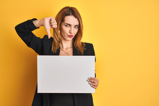 Redhead business caucasian woman holding banner over yellow isolated background with angry face, negative sign showing dislike with thumbs down, rejection concept