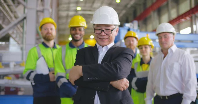 Asian businessman smile face portrait. Korean business person in formal wear, glasses and hardhat looks to camera crosses arms over chest. On background staff team employees at modern factory facility