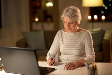 technology, old age and people concept - senior woman writing to notebook with laptop at home in...