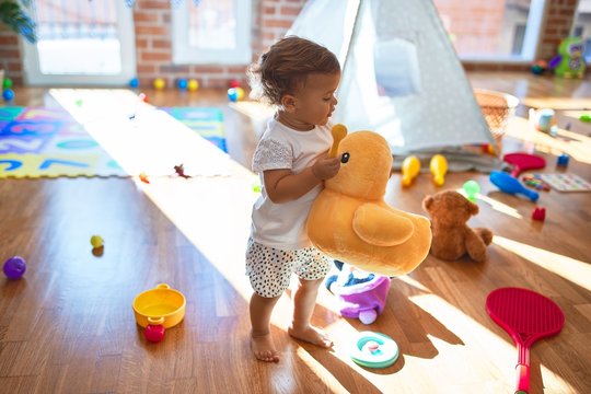 Adorable toddler holding duck doll standing around lots of toys at kindergarten