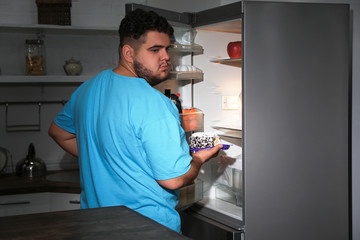 Fototapeta premium Depressed overweight man taking cake out of refrigerator at night