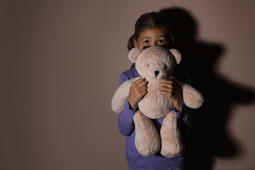 Sad little girl with teddy bear near beige wall, space for text. Domestic violence concept
