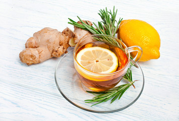 Tea in a glass cup with the ginger roots, rosemary branches and lemon on a white wooden background.