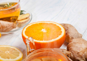 Tea in teapot and cup of tea with ginger roots, rosemary, orange and lemon on white wooden background. Selective focus.