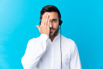 Telemarketer man working with a headset over isolated blue background covering a eye by hand