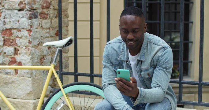 Cheerful African American Young Man Having a Video Call via his Smartphone. Guy Waving and Smiling while Sitting near Stylish Bike with Old Metal Fance at the Background.