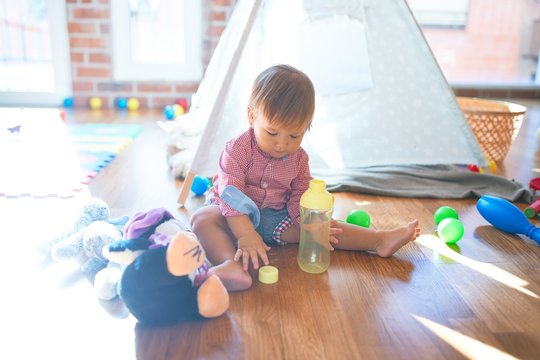 Adorable toddler holding feeding bottle around lots of toys at kindergarten