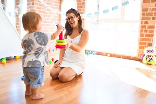 Beautiful teacher and toddler building pyramid using hoops around lots of toys at kindergarten