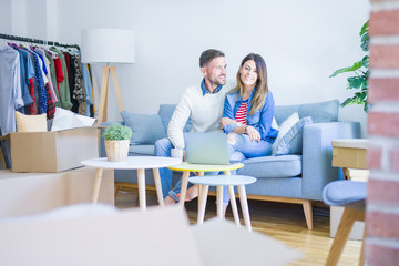 Young beautiful couple sitting on the sofa drinking cup of coffee using laptop at new home around cardboard boxes