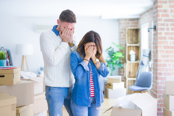 Young beautiful couple standing at new home around cardboard boxes with sad expression covering face with hands while crying. Depression concept.