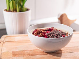 Eine weiße Keramikschüssel mit Joghurt, Pecannüssen, Kakaonibs und Himbeeren, steht in der Küche auf einem hellen Holzbrett.
