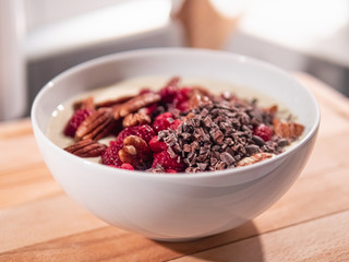 Eine weiße Keramikschüssel mit Joghurt, Pecannüssen, Kakaonibs und Himbeeren, steht in der Küche auf einem hellen Holzbrett.