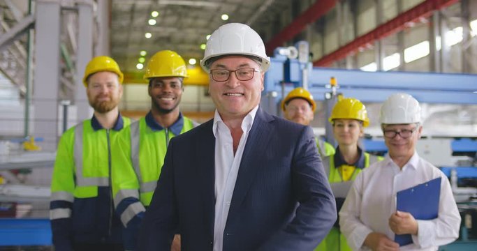 Caucasian businessman smile face portrait. Positive smiling business person in formal wear, helmet looking to camera crosses arms over chest. On background staff team employees at heavy industry plant
