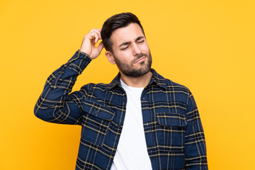 Young handsome man with beard over isolated yellow background having doubts while scratching head