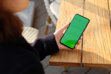 over shoulder of one woman holding green screen phone on wooden table under sunshine. blur background