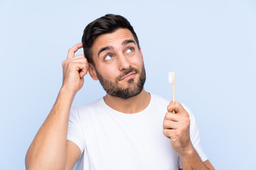 Young handsome man with beard brushing his teeth over isolated background having doubts and with...