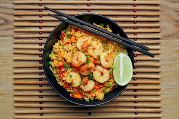 Traditional shrimps stir fried with eggs, vegetables and rice in a black bowl on a bamboo placemat. Healthy Asian meal with sesame and lime. Top view, directly above shot.