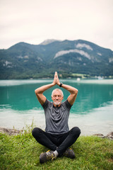 A senior man pensioner sitting by lake in nature, doing yoga exercise.