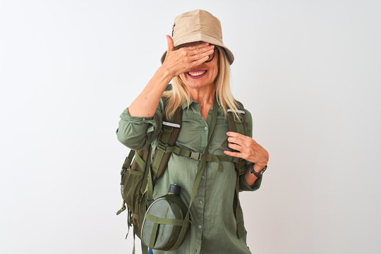 Middle age hiker woman wearing backpack hat canteen glasses over isolated white background smiling and laughing with hand on face covering eyes for surprise. Blind concept.