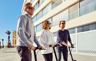 Fototapeta na wymiar Three best friends young 20s -30s girl and guys spend time outdoors gathered together driving on electric scooter modern land vehicle, easy comfort usage, technology urban transportation concept image