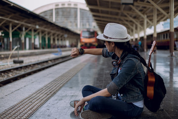 Fototapeta na wymiar Outdoors lifestyle of young women. tourist girl wearing hat with backpack and looking at her wrist watch while sitting on floor and waiting for the train at the railway station, Travel concept.