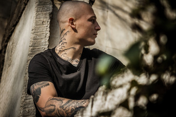 Young attractive stern guy in tattoos sits on a window frame of a destroyed building. Brutal young...