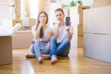 Beautiful couple sitting on the floor holding smartphone at new home around cardboard boxes serious face thinking about question, very confused idea