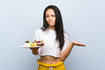 Teenager asian girl holding lots of different mini cakes making doubts gesture while lifting the shoulders