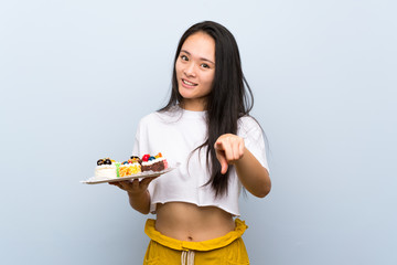 Teenager asian girl holding lots of different mini cakes points finger at you with a confident expression