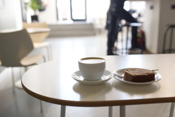 Cappuccino in a white mug with a piece of cake on a white table in a cafe close-up.