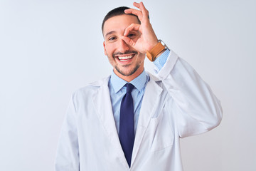 Young doctor man wearing white coat over isolated background doing ok gesture with hand smiling, eye looking through fingers with happy face.